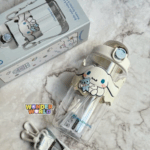 A clear plastic water bottle featuring a cream-colored cap with a light blue pop-up button, a soft silicone protective sleeve showing Cinnamoroll holding a blue teddy bear, and an adjustable matching character shoulder strap lying nearby next to its original product packaging