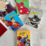 An array of four miniature spiral pocket notebooks featuring classic DC Comics heroes, showcase-styled with bright leaf-green, sky-blue, stone-grey, and warm ochre-yellow covers highlighting retro comic-book illustrations of The Flash, Superman, Batman, and the Justice League