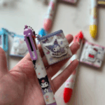 A hand holding a light purple multi-color shuttle pen decorated with Kuromi and star prints, connected by a purple ball chain to a tiny, matching square pocket notebook charm with a puffy Kuromi character clasp