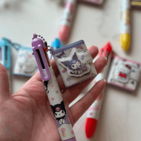 A hand holding a light purple multi-color shuttle pen decorated with Kuromi and star prints, connected by a purple ball chain to a tiny, matching square pocket notebook charm with a puffy Kuromi character clasp