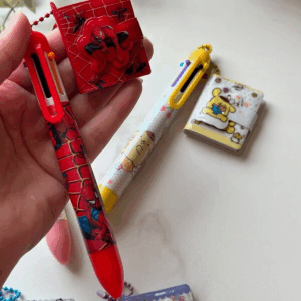 A hand holding a vibrant red multi-color shuttle pen with Spider-Man graphics from the file WW398-600x600.jpeg, connected by a red ball chain to a tiny square web-patterned pocket notebook with a puffy Spider-Man clasp
