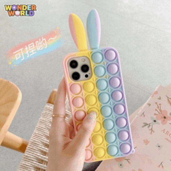 A close-up display of a whimsical silicone bunny phone case held in a hand, featuring vertical stripes of soft pop-it bubbles in a soothing gradient of pastel pink, sunlit yellow, mint blue, and lavender purple, crowned by two playful, upright 3D bunny ears