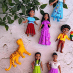 A flat-lay arrangement of seven beautifully sculpted play figurines on a white textured background, featuring beloved characters in vibrant dresses of lilac, magenta, cerulean blue, and lime green, displayed alongside a spotted yellow jaguar and leafy green vines