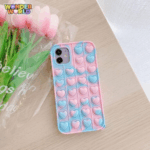 A close-up display of a premium protective phone case resting on a light wooden table next to a bouquet of soft pink tulips, showcasing an organic marbled blend of pastel sky blue and blossom pink with rows of responsive pop-it bubbles sculpted into sweet miniature heart silhouettes