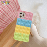 A close-up display of a premium protective phone case resting on a light wooden stool next to a cozy pink-and-white checkered backdrop, showcasing a horizontal color-block gradient of soft blossom pink, pastel sky blue, sunny buttercup yellow, and gentle mint green with rows of responsive pop-it bubbles sculpted into sweet miniature heart silhouettes