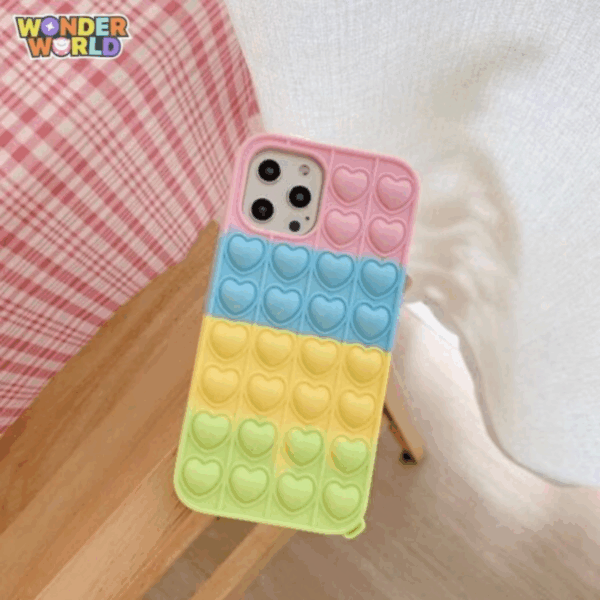 A close-up display of a premium protective phone case resting on a light wooden stool next to a cozy pink-and-white checkered backdrop, showcasing a horizontal color-block gradient of soft blossom pink, pastel sky blue, sunny buttercup yellow, and gentle mint green with rows of responsive pop-it bubbles sculpted into sweet miniature heart silhouettes