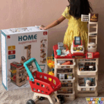 A multi-tiered toy supermarket checkout stand in cream and crimson red with shelves stocked with play goods, a toy register, and a small red shopping cart with a turquoise handle
