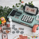 A high-end interactive toy checkout terminal in soft sage green, featuring a mock smart display screen, cream-colored buttons, and a warm terracotta barcode scanner, displayed alongside play money, a miniature credit card, and tiny grocery replicas