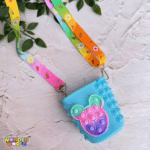 A close-up display of a whimsical cup-shaped silicone pop-it purse in soft sky blue, featuring a front-facing pop-it panel sculpted into an adorable miniature mouse-head silhouette with a vibrant mint green, pink, and purple gradient, complete with a colorful patterned shoulder strap decorated with tiny sweet treats