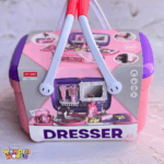 A structured play vanity basket featuring a soft pastel pink base, a rich purple lid, and dual white swing handles with vibrant coral grips, shown with its illustrated packaging wrap detailing the pretend makeup tools inside