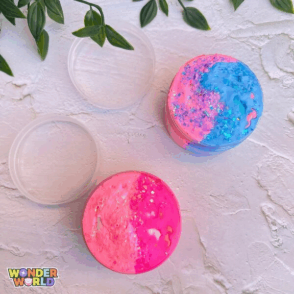 A top-down view of two round clear containers filled with vibrant, glittering sensory slimes on a rustic white textured surface with soft green leaves in the corner; one container showcases a beautiful dual-tone swirl of hot pink and soft baby pink dusted with sparkling star glitters, while the other features a striking split of neon pink and sky blue topped with shimmering iridescent flakes