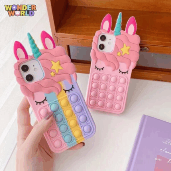 A close-up display of two whimsical silicone unicorn phone cases set against a warm wooden backdrop, one featuring a solid sweet-pink design and the other a pastel rainbow gradient of pink, blue, yellow, and lavender, both showcasing sculpted sleeping eyes, a mint-green horn, and a responsive pop-it bubble panel