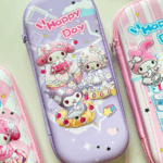 A close-up view of a protective, hard-shell zippered pencil case in soft pastel lavender, featuring My Melody, Kuromi, and My Sweet Piano dressed in maid outfits while resting on giant waffles and fruit cupcakes under the pink-and-white cursive script "Happy Day", shown between two other pink-themed Sanrio pencil cases