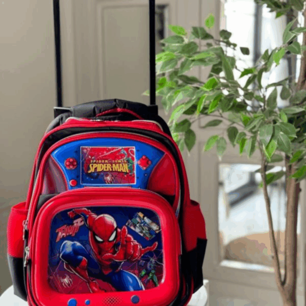 A premium rolling school backpack in rich crimson red and deep royal blue, featuring a glossy upper panel styled like a classic handheld gaming console and a lower glossy 3D pocket showing Spider-Man mid-action, set against a sleek black canvas body with a telescoping handle
