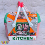A structured play kitchen basket featuring a soft rose-pink base, a contrasting jade-green lid, and dual curved white handles topped with vibrant coral-red hand grips, shown packaged in an illustrated sleeve