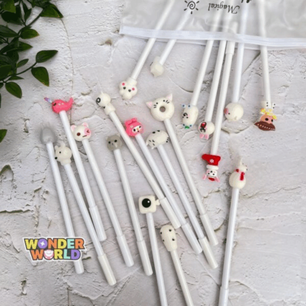 A flat-lay view of several slender cream-white gel pens spilling out of a frosted slide-zip pouch labeled "Magical" on a rustic white textured surface with soft green leaves in the corner, each pen crowned with a unique, colorful 3D silicone topper including a winking pink narwhal, a round cat-donut, a grey kitten, a festive red stocking, and a cozy snowman