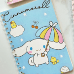 A soft sky-blue spiral notebook featuring an illustration of Cinnamoroll holding a striped pastel umbrella, surrounded by little yellow bird friends, floating cherries, a white cloud, and scattering white stars, with "Cinnamoroll" written in a black cursive font across the top left