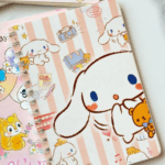 A wire-bound notebook featuring a warm peach and cream vertical striped background with various sketched illustrations of Cinnamoroll, a travel suitcase, a retro camera, a red double-decker bus, and a little brown teddy bear